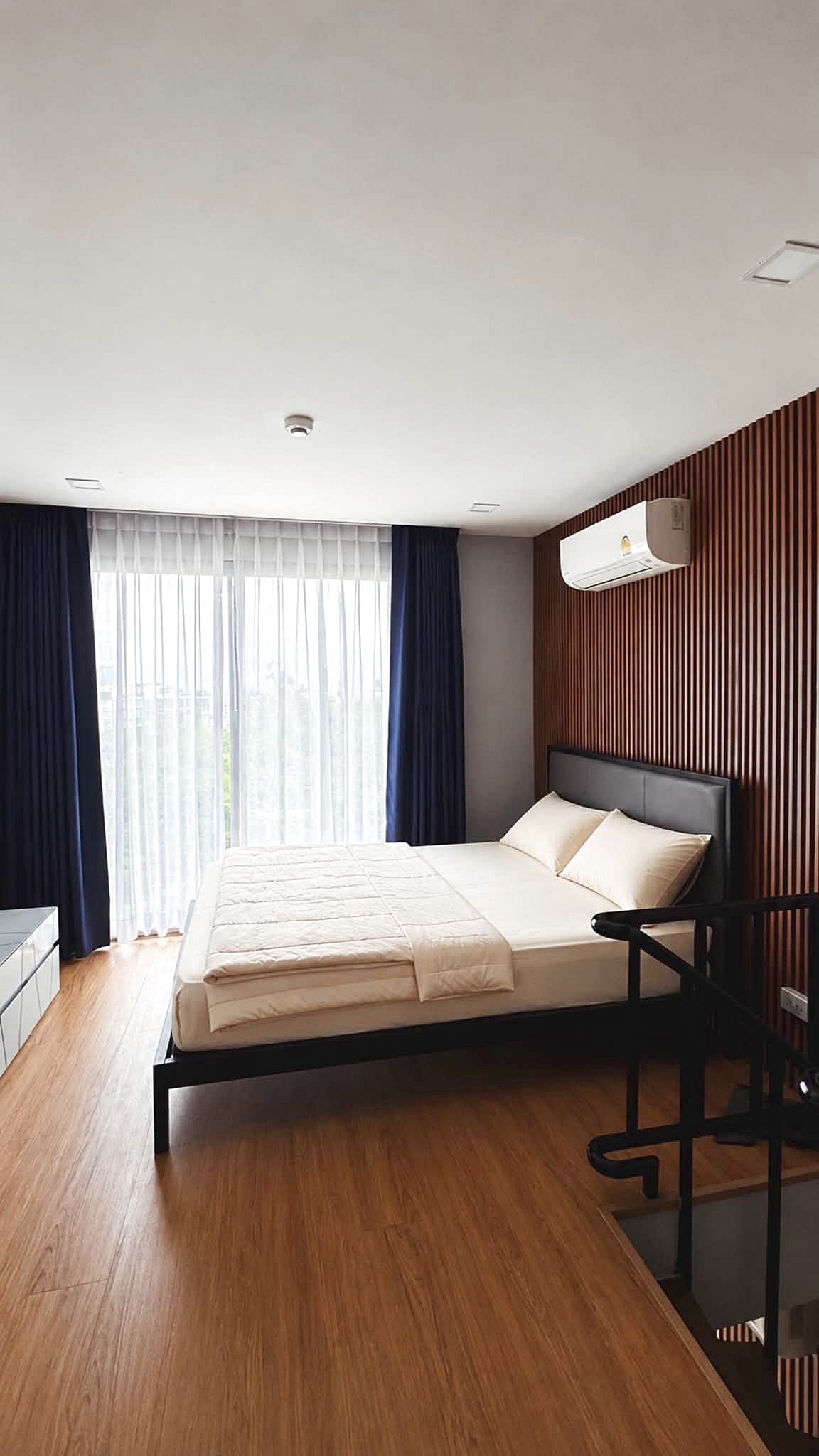 Laguna_2_Pattaya_Sale_Dlc644_3 Hotel bedroom with a low dark wood bed, beige linens, and a vertical wood slat accent wall; large window with sheer white curtains and navy drapes, wood floor, and wall-mounted AC unit.