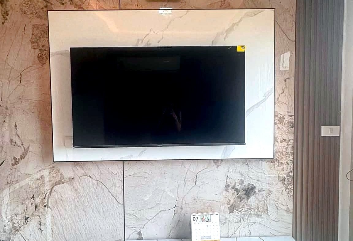Wall-mounted TV on a marble panel with a square clock above; white modern media console and a remote on a white tiled table in a bright living room.