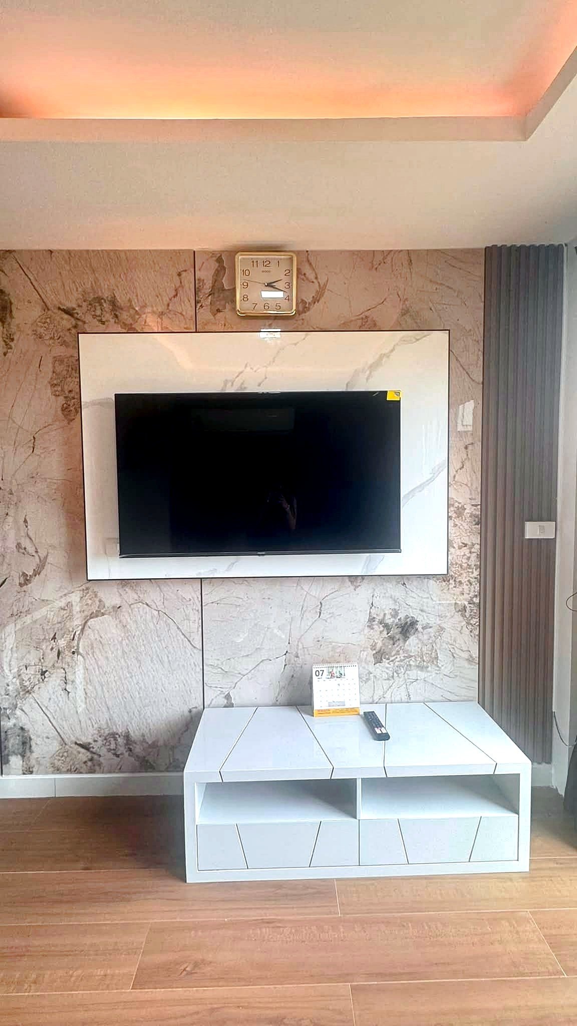 Laguna_2_Pattaya_Sale_Dlc644_5 Wall-mounted TV on a marble panel with a square clock above; white modern media console and a remote on a white tiled table in a bright living room.