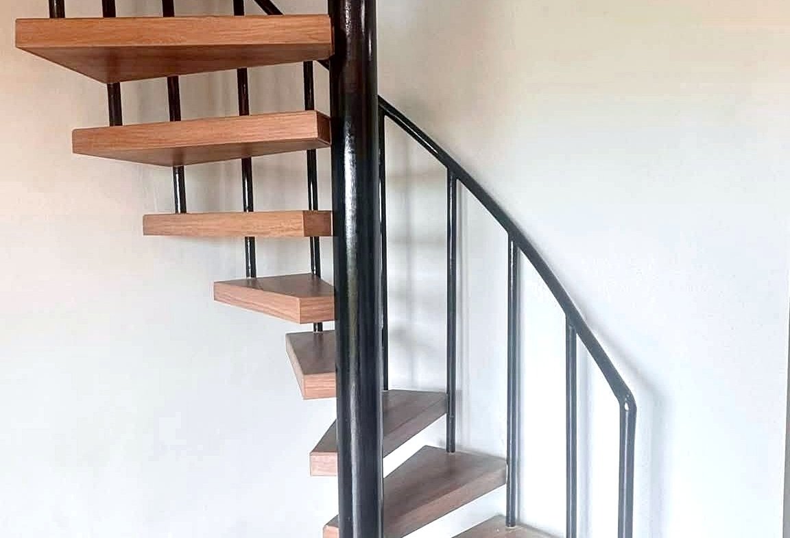 Spiral staircase with wooden steps and black metal railing winding around a central black pillar in a bright interior.