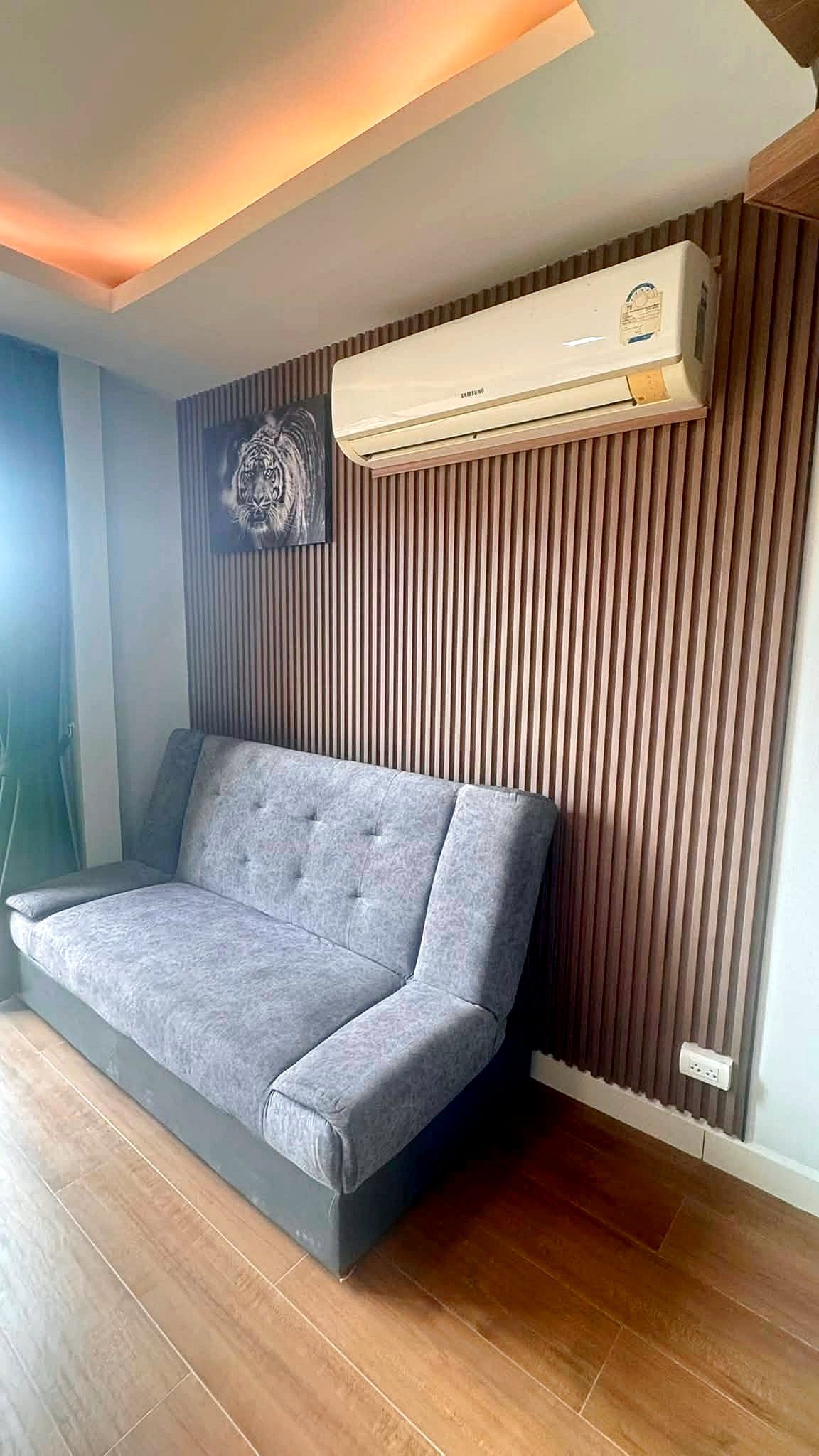 Laguna_2_Pattaya_Sale_Dlc644_7 Gray fabric sofa against a vertical wood-slatted wall, with a white wall AC unit above and a tiger artwork nearby.