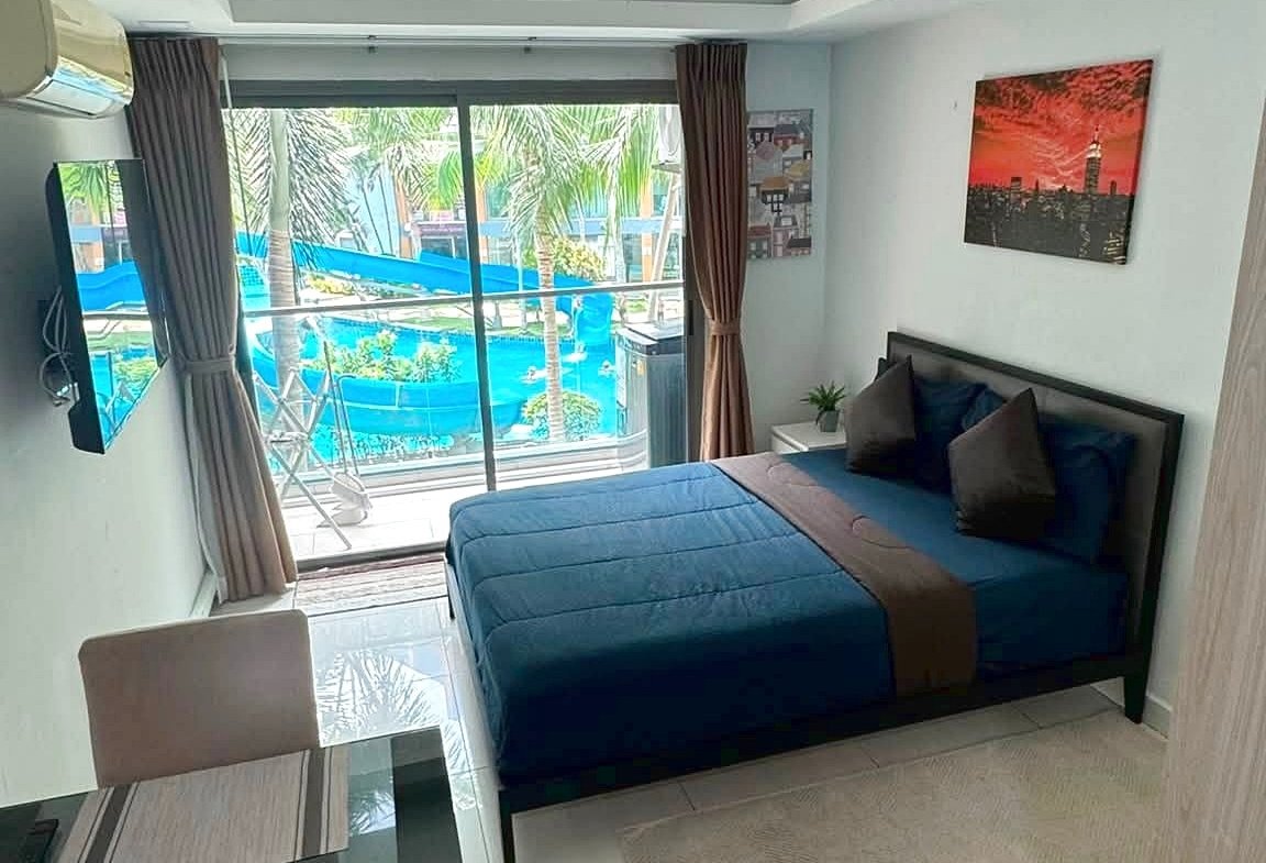 Hotel room with a blue bedspread facing a sliding glass door to a balcony; pool and palm trees visible outside.