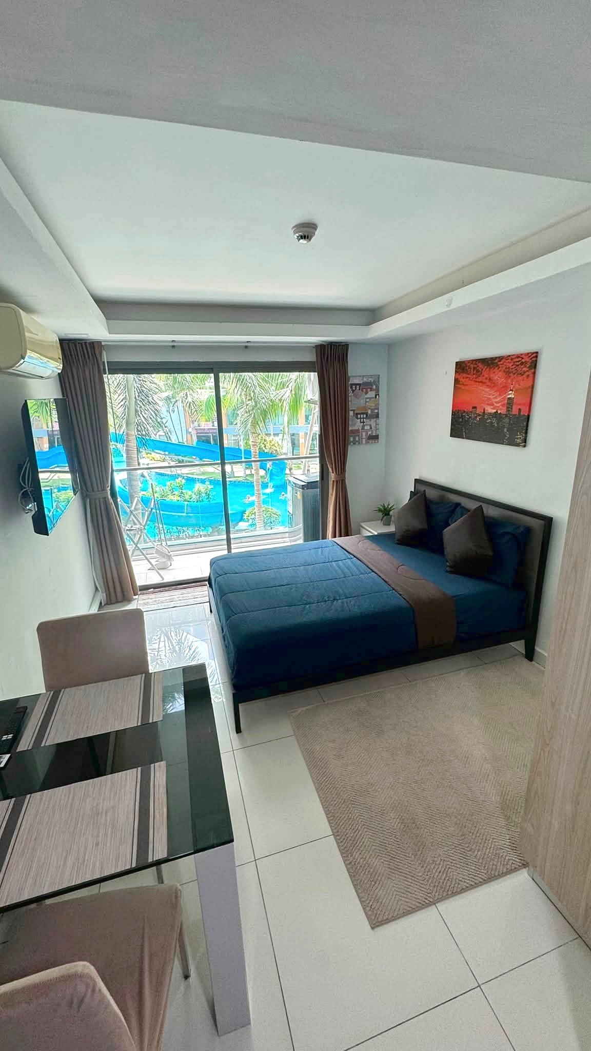 Hotel room with a blue bedspread facing a sliding glass door to a balcony; pool and palm trees visible outside.