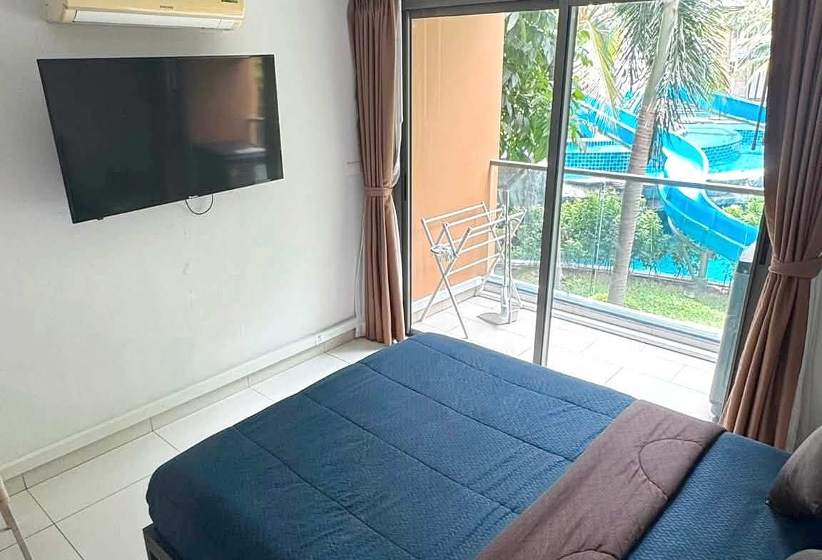 Bedroom with a wall-mounted TV and a ceiling air conditioner, bed in the foreground, sliding glass door to a balcony with a pool and water slide beyond.