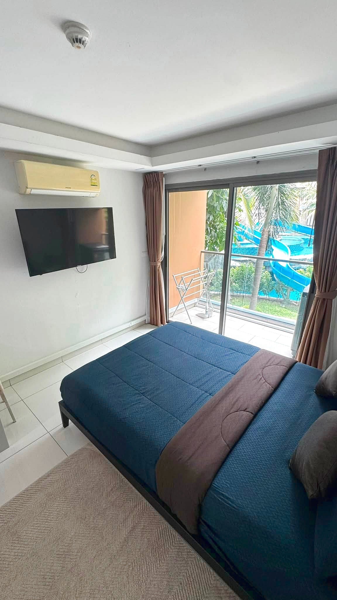 Bedroom with a wall-mounted TV and a ceiling air conditioner, bed in the foreground, sliding glass door to a balcony with a pool and water slide beyond.