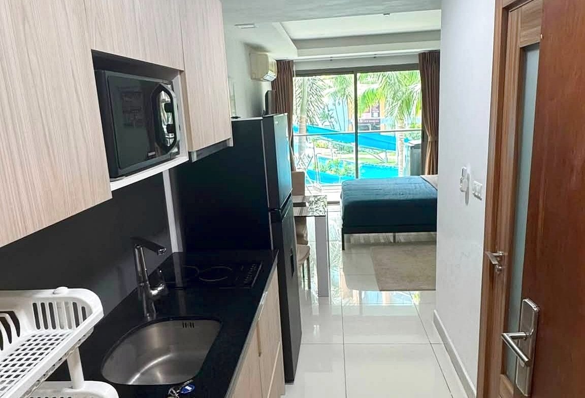Narrow kitchen with light wood cabinets and black countertop, sink and dish rack, opening to a living area with a bed/sofa and sliding doors to a pool outside.