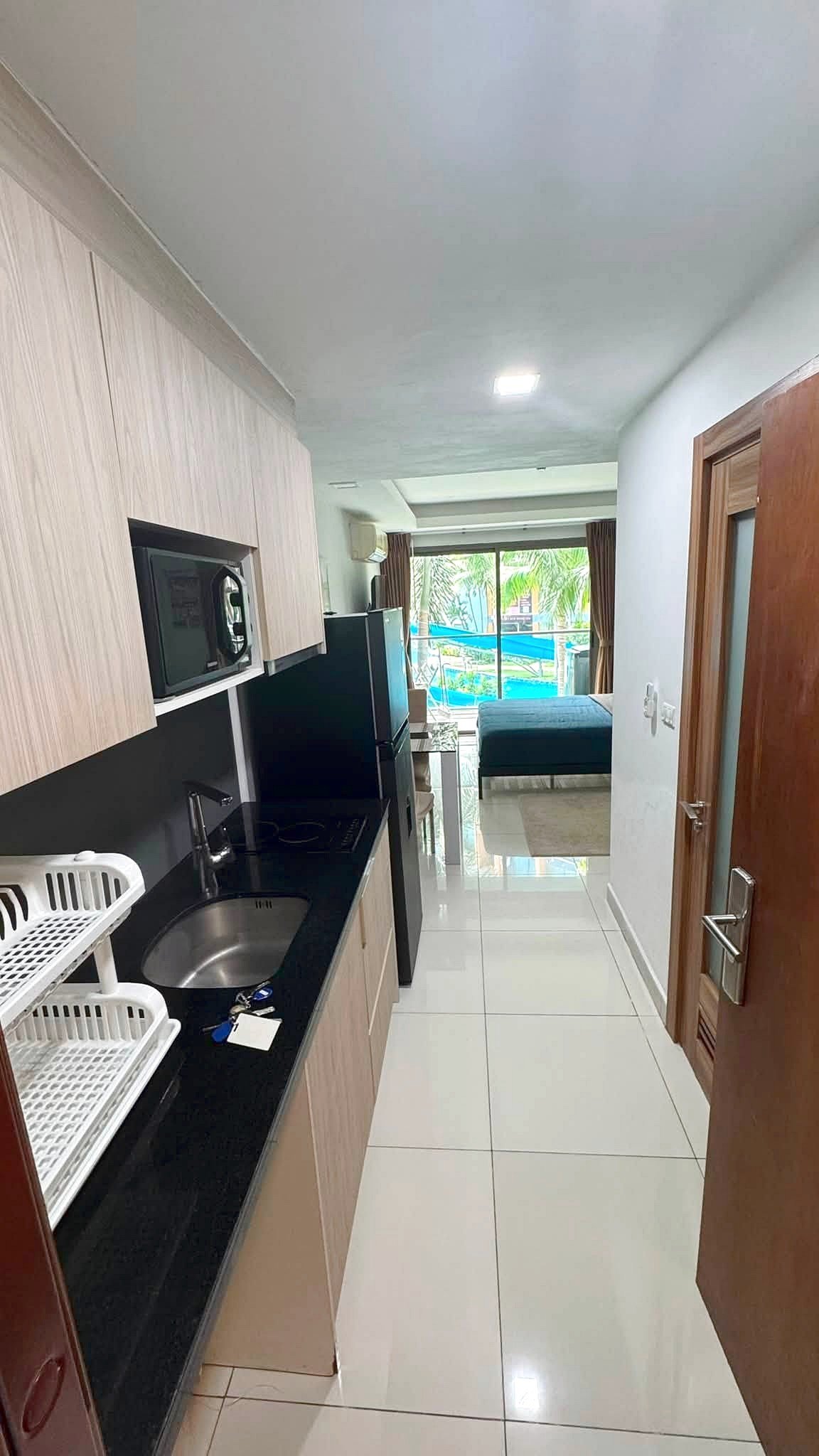 Narrow kitchen with light wood cabinets and black countertop, sink and dish rack, opening to a living area with a bed/sofa and sliding doors to a pool outside.
