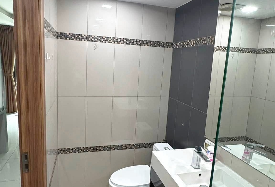 Modern bathroom with white and gray tiled walls, a glass shower partition, and a white vanity with sink and mirror. A toilet sits beside the vanity; decorative tile strip runs horizontally along the walls.