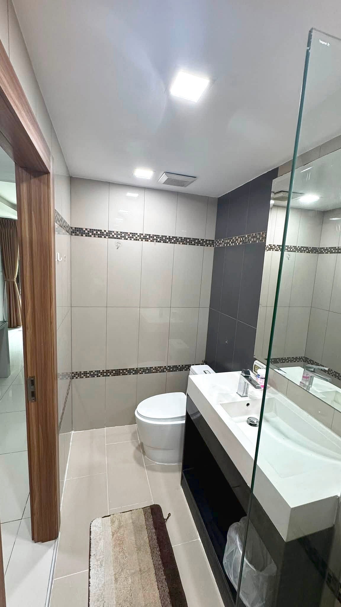 Modern bathroom with white and gray tiled walls, a glass shower partition, and a white vanity with sink and mirror. A toilet sits beside the vanity; decorative tile strip runs horizontally along the walls.