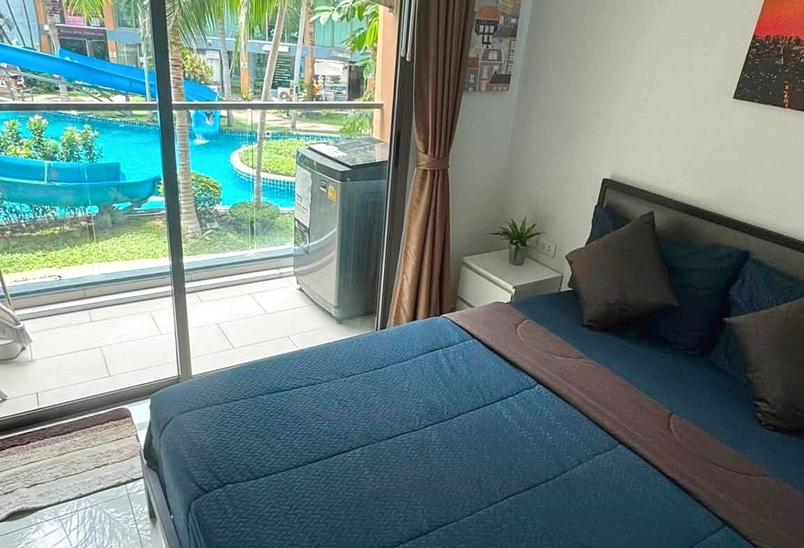 Bedroom with a blue bedspread and multiple pillows, sliding glass door to a balcony with a pool and palm trees outside, brown curtains drawn to the side.
