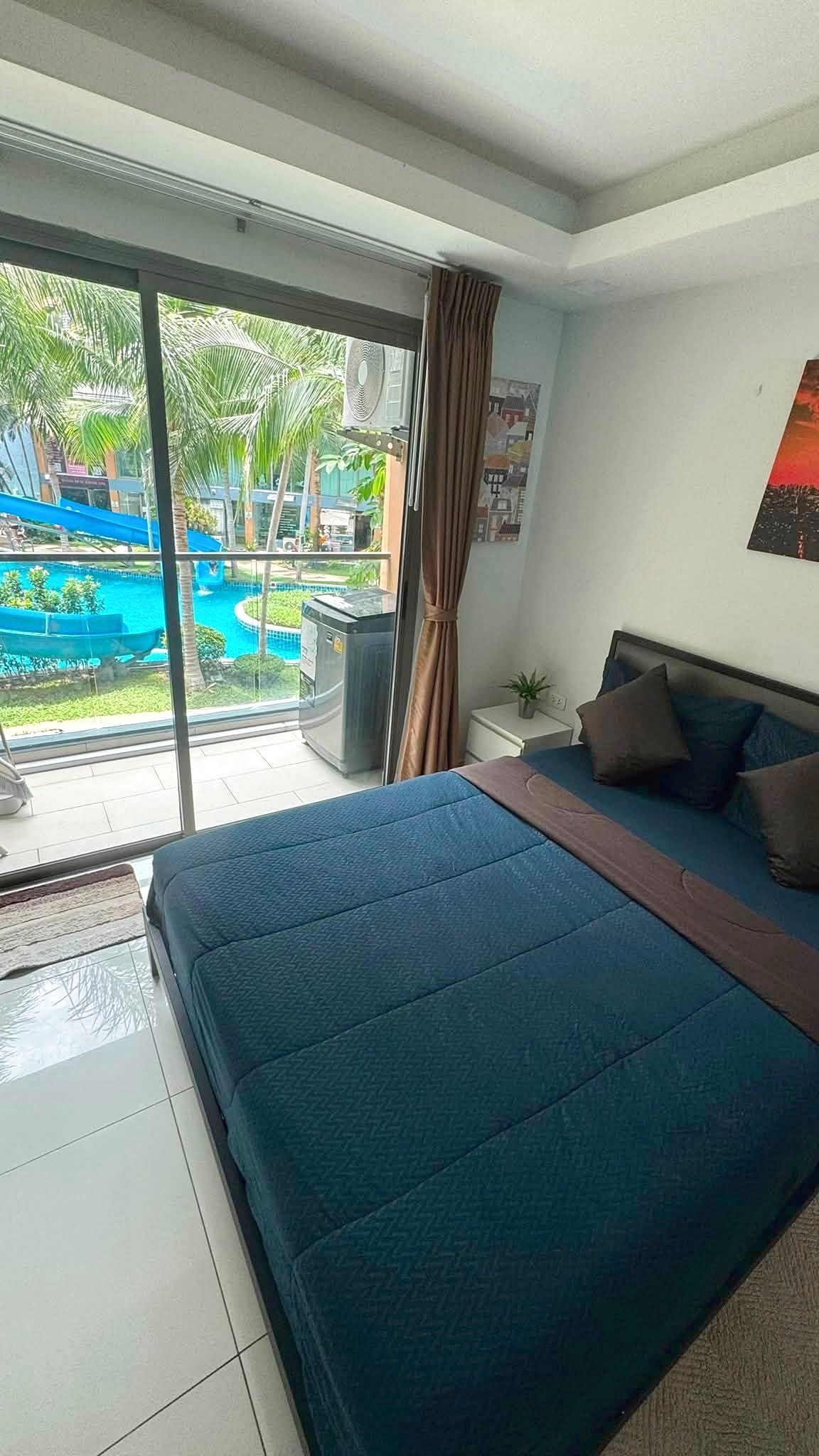 Bedroom with a blue bedspread and multiple pillows, sliding glass door to a balcony with a pool and palm trees outside, brown curtains drawn to the side.