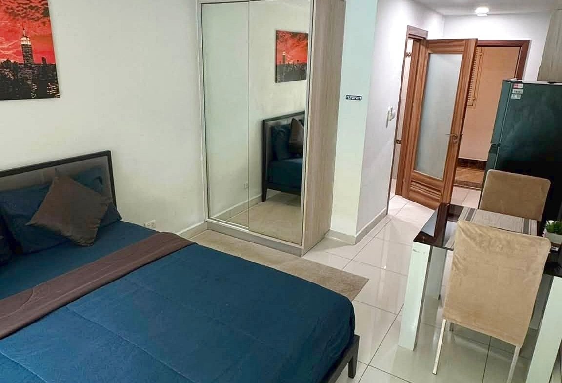 Modern hotel bedroom with a blue duvet, a low bed frame, and a mirrored wardrobe beside it. A glass dining table with beige chairs sits nearby, and an open wooden door leads to a hallway.
