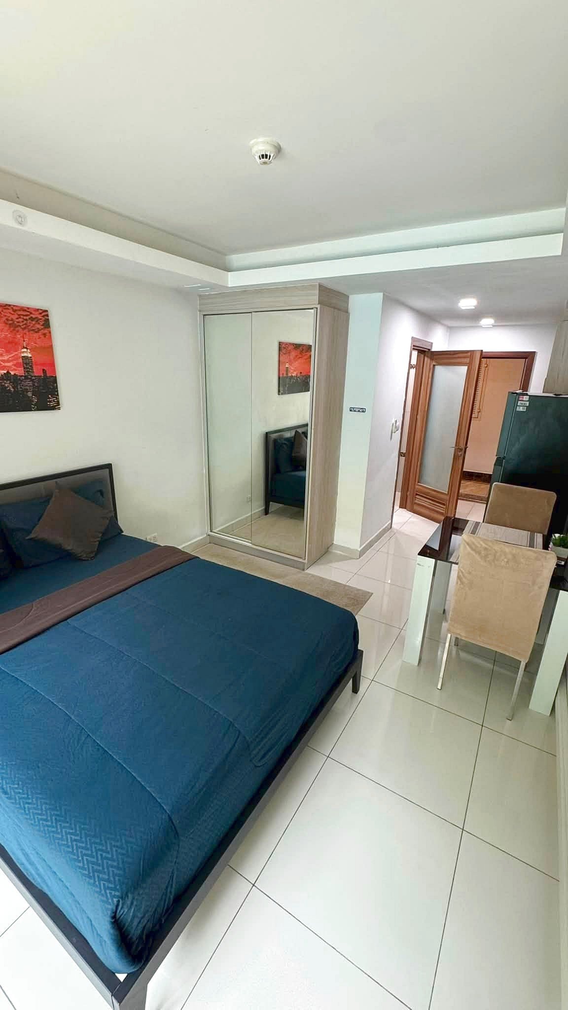 Modern hotel bedroom with a blue duvet, a low bed frame, and a mirrored wardrobe beside it. A glass dining table with beige chairs sits nearby, and an open wooden door leads to a hallway.
