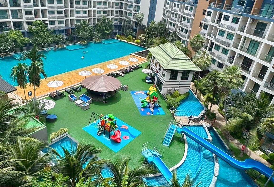 Resort pool area surrounded by multi-story buildings, palm trees, and colorful kids' playground equipment including blue slides and shaded lounging areas.