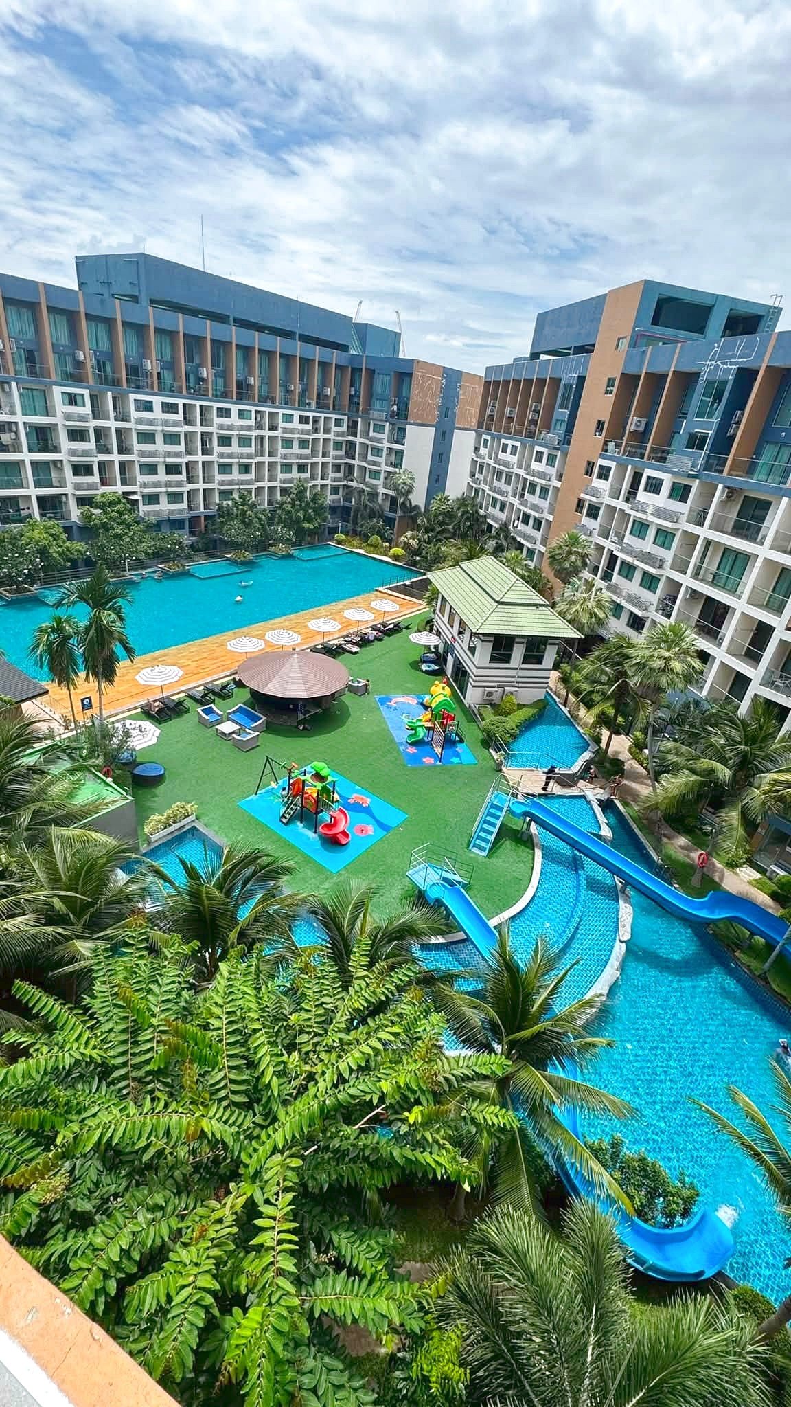 Resort pool area surrounded by multi-story buildings, palm trees, and colorful kids' playground equipment including blue slides and shaded lounging areas.