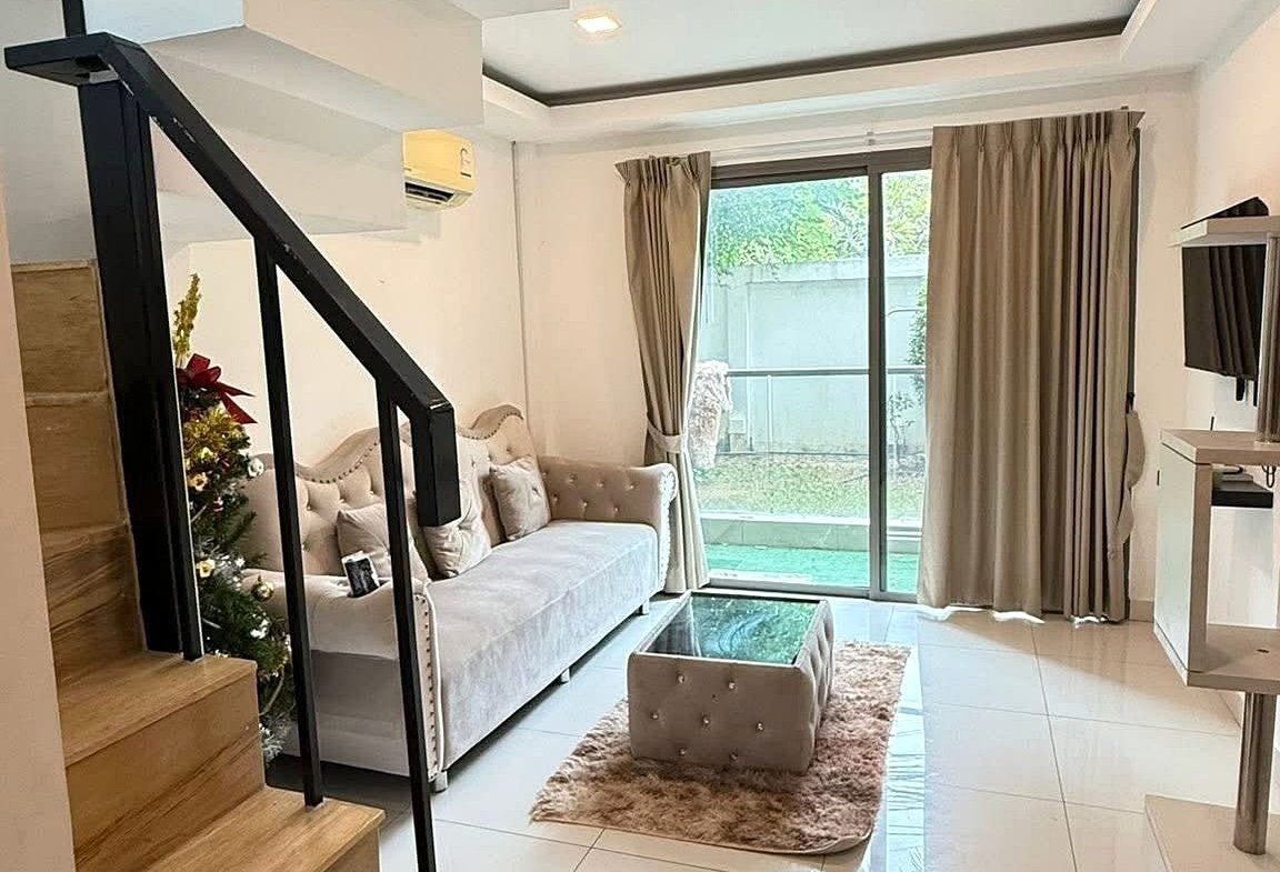 Bright modern living room with white tiled floor, beige sofa, glass coffee table, and a small rug under the seating area beside a staircase.