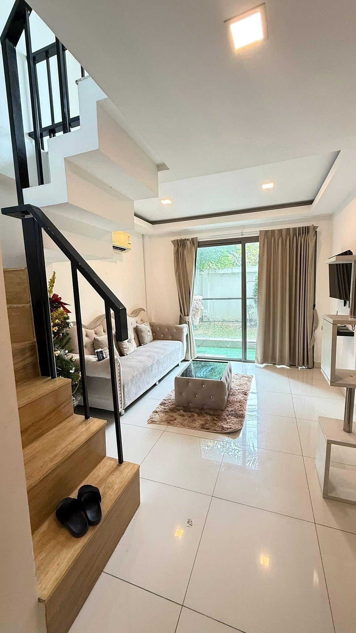 Laguna_Beach_2_Pattaya_Dlc627_2 Bright modern living room with white tiled floor, beige sofa, glass coffee table, and a small rug under the seating area beside a staircase.