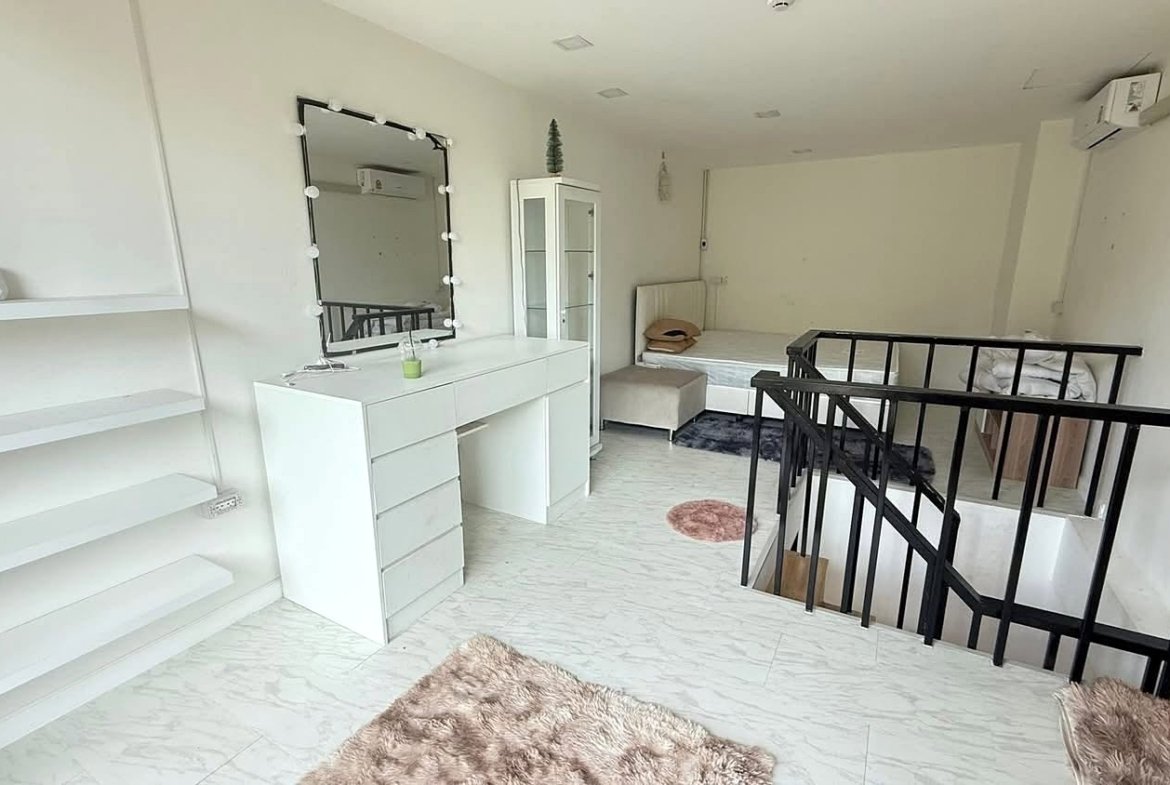 Bright white loft room with a makeup vanity, mirror framed with bulbs, and a metal railing nearby.