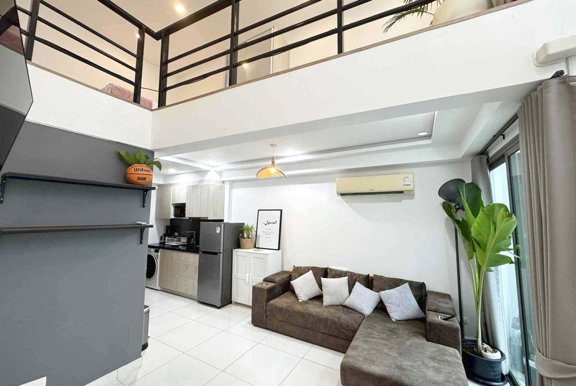 Modern open-plan living area: gray kitchen island, stainless steel appliances, white tile floor, and a brown sectional sofa with white cushions.
