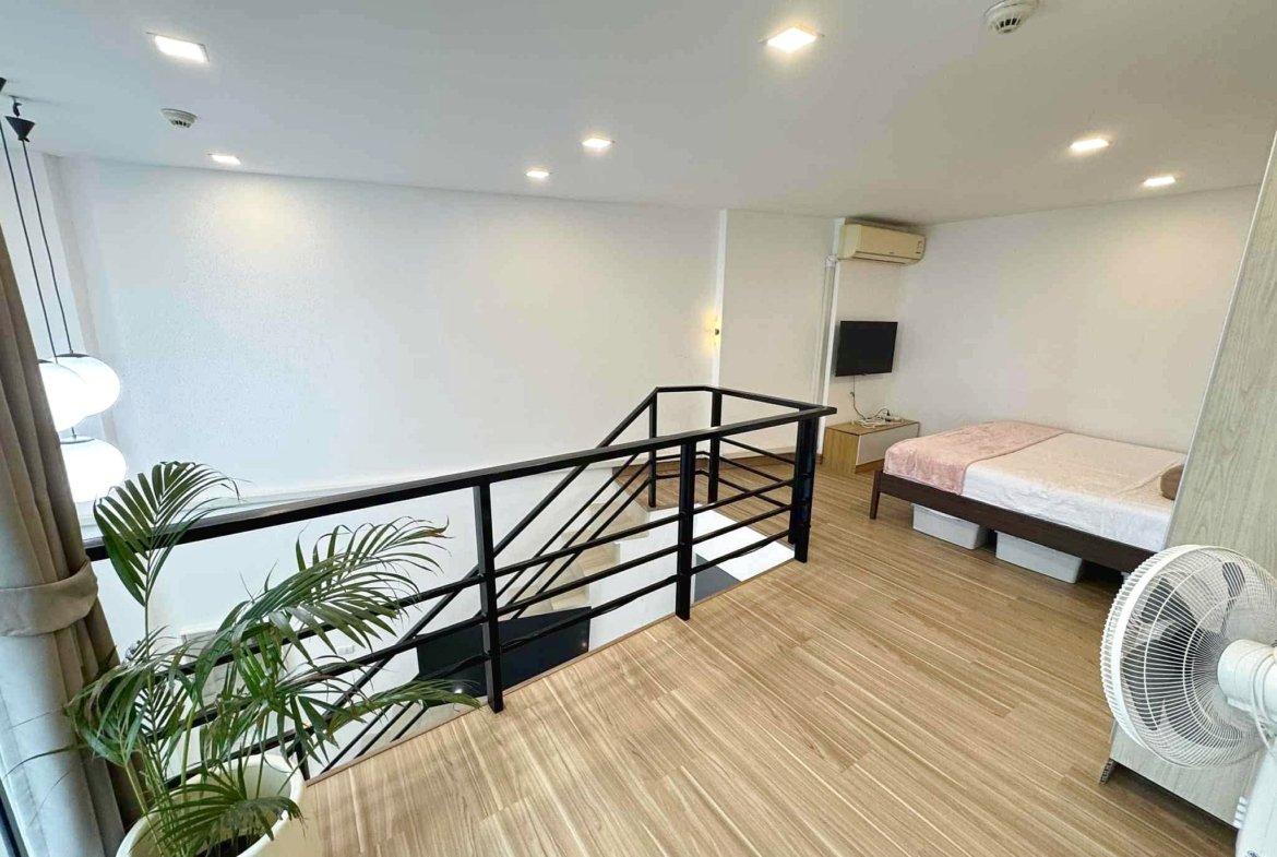 Bright loft bedroom with two beds, wall-mounted TV, ceiling lights, and an AC unit; plants near the railing.