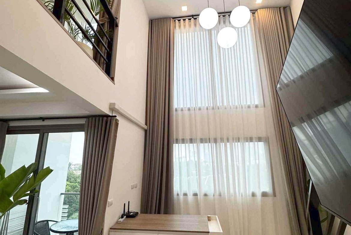 Bright two-story living area with large windows and sheer curtains, featuring hanging globe lights and a balcony with outdoor seating visible outside.
