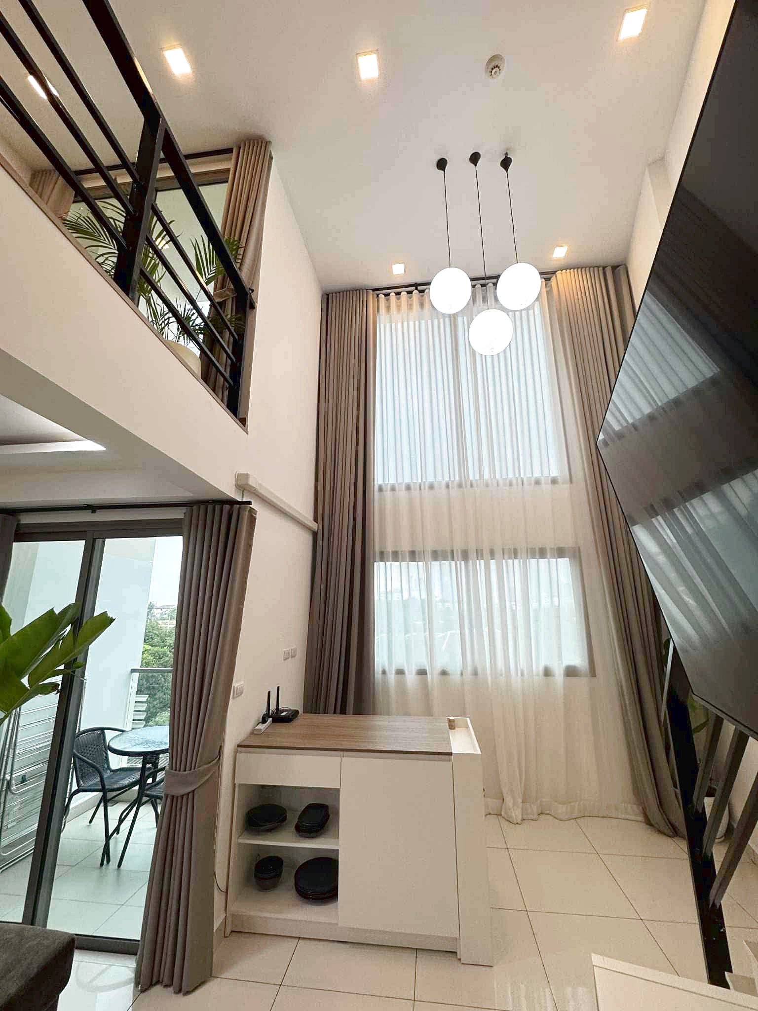 Laguna_Beach_2_Rent_Rdl362_4 Bright two-story living area with large windows and sheer curtains, featuring hanging globe lights and a balcony with outdoor seating visible outside.