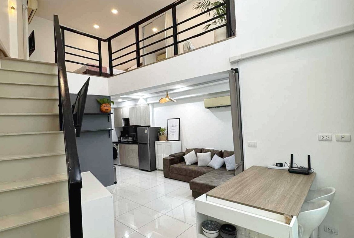 Open-concept living space with white tiled floor, stairs to a mezzanine, and black railing overlooking a compact kitchen and sofa area.