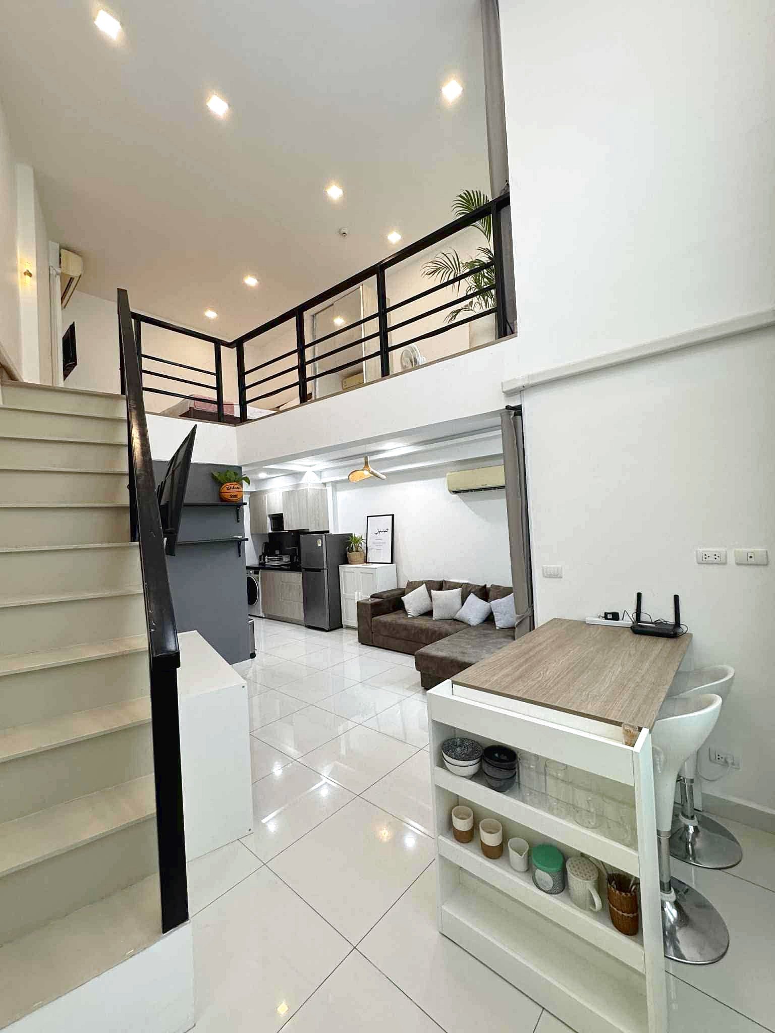 Laguna_Beach_2_Rent_Rdl362_5 Open-concept living space with white tiled floor, stairs to a mezzanine, and black railing overlooking a compact kitchen and sofa area.