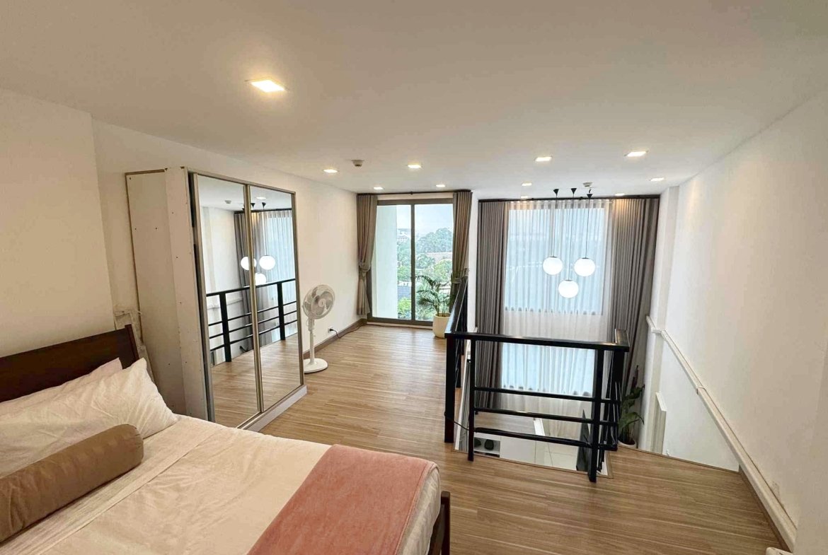 Loft bedroom with two single beds in foreground, wooden floor, and glass railing; large windows with sheer curtains in back.
