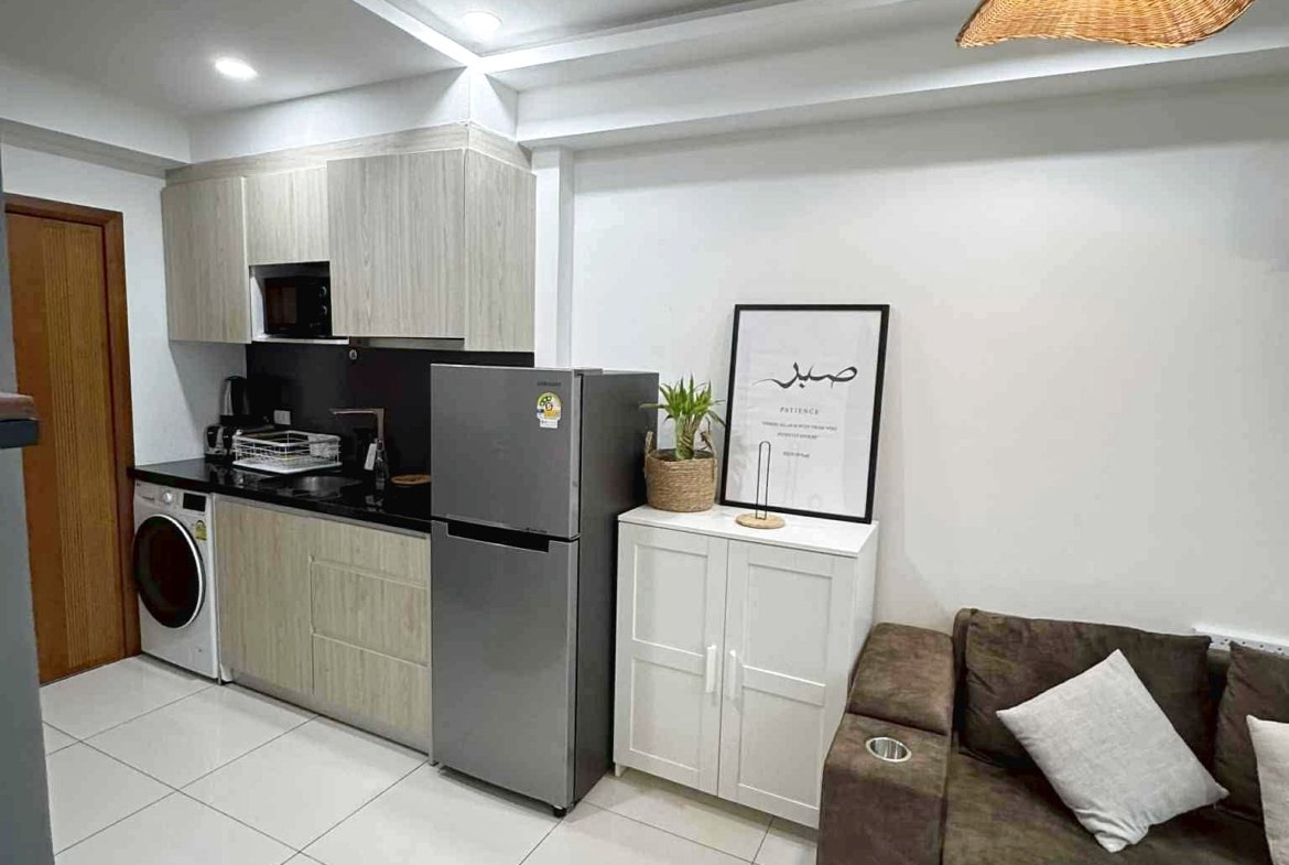 Open-plan kitchen with light wood cabinets, stainless fridge and washing machine, white tiled floor, and a brown sofa with pillows nearby.