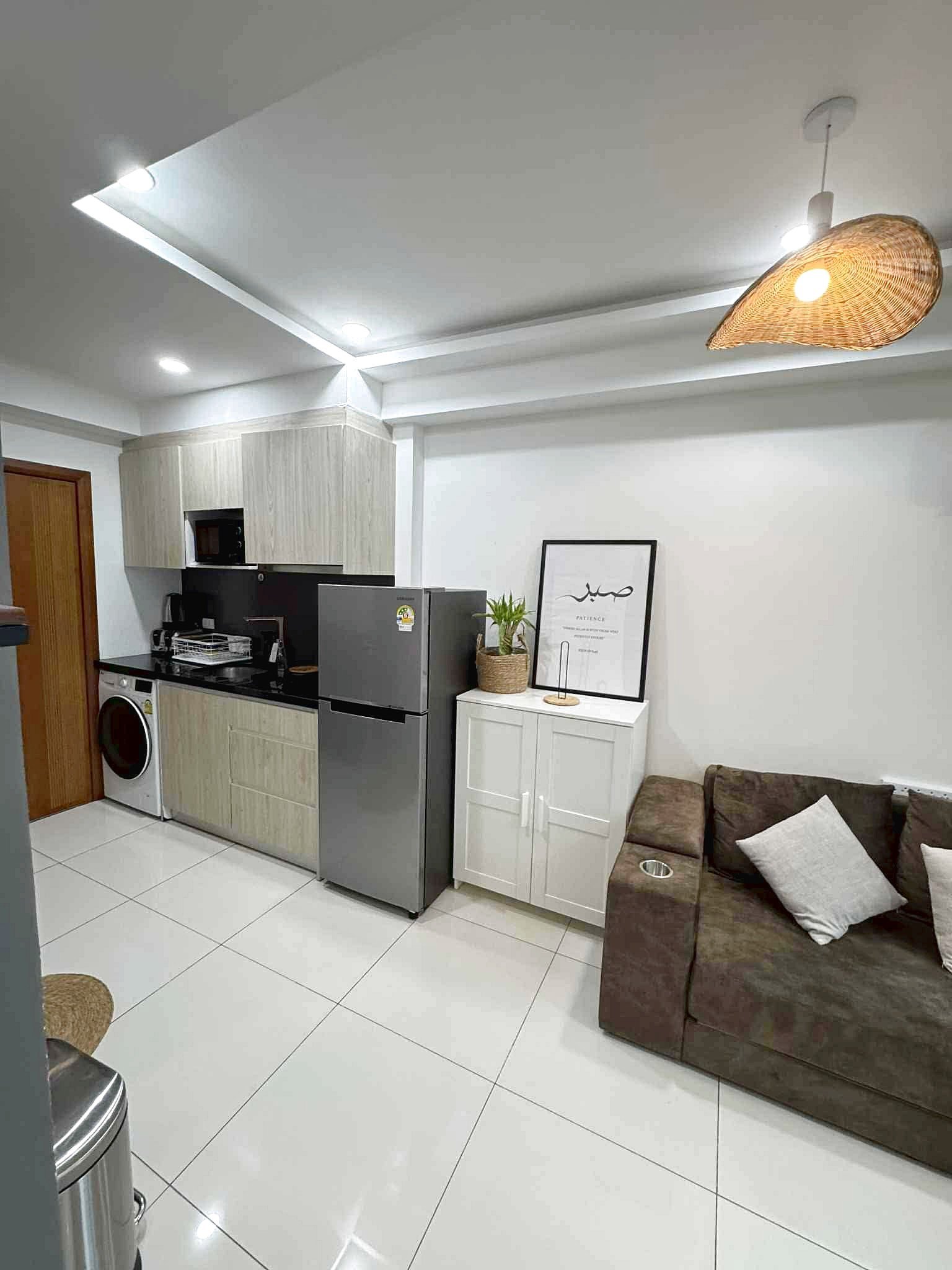 Laguna_Beach_2_Rent_Rdl362_7 Open-plan kitchen with light wood cabinets, stainless fridge and washing machine, white tiled floor, and a brown sofa with pillows nearby.