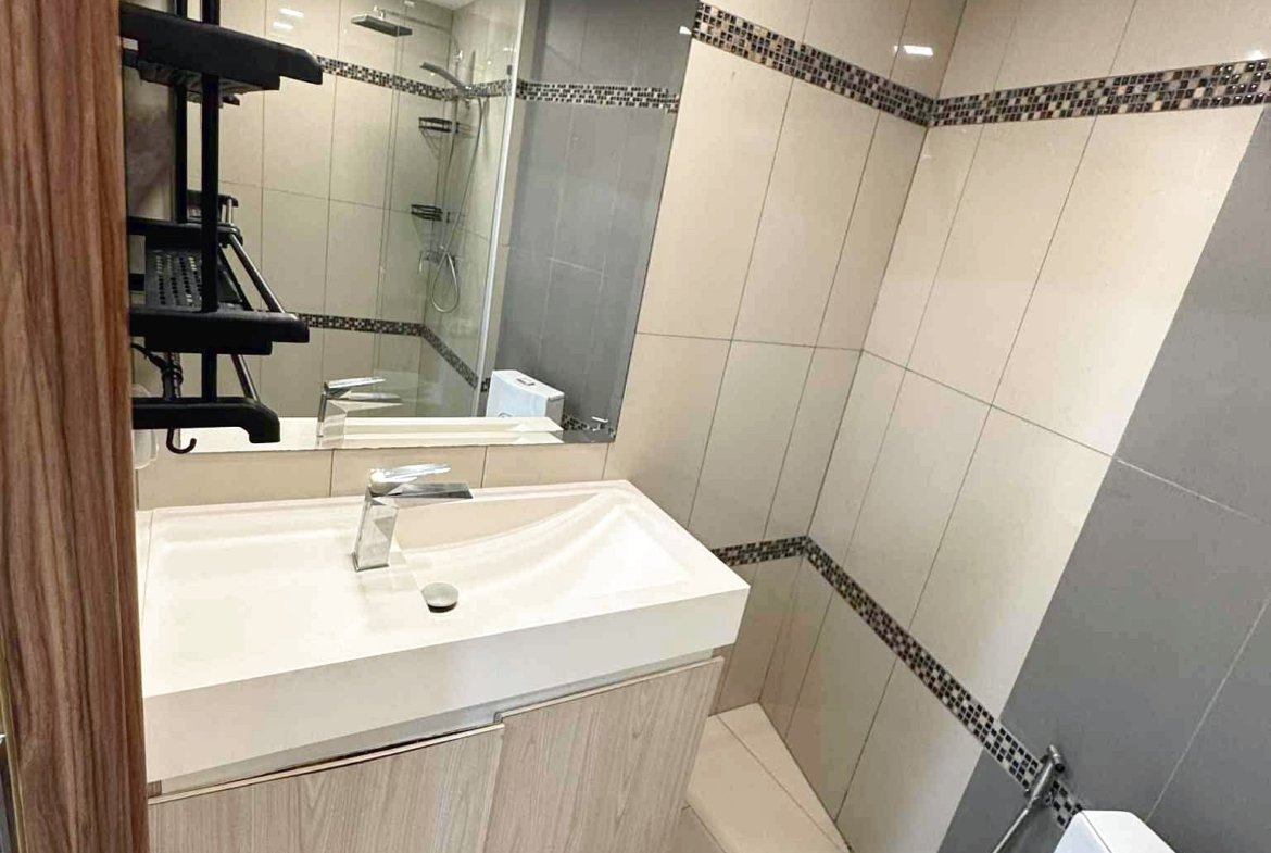 Modern bathroom with a white sink vanity, large wall mirror, beige tiled walls, and a glass-enclosed shower; washing machine visible nearby.