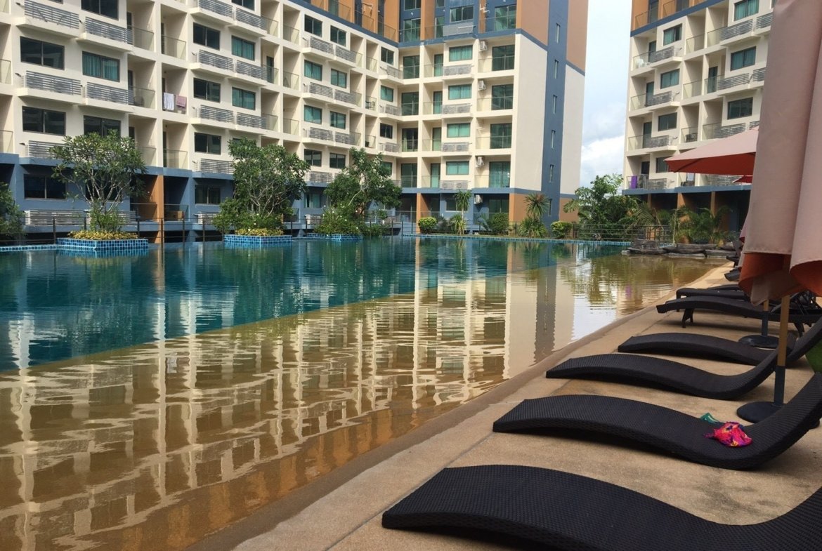 Outdoor hotel pool area with modern low-rise buildings, lounge chairs, and umbrellas along the pool deck.