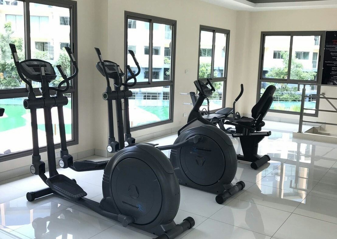 Bright gym room with cardio machines: two ellipticals, two stationary bikes, and a treadmill by large windows and white tiled floor.