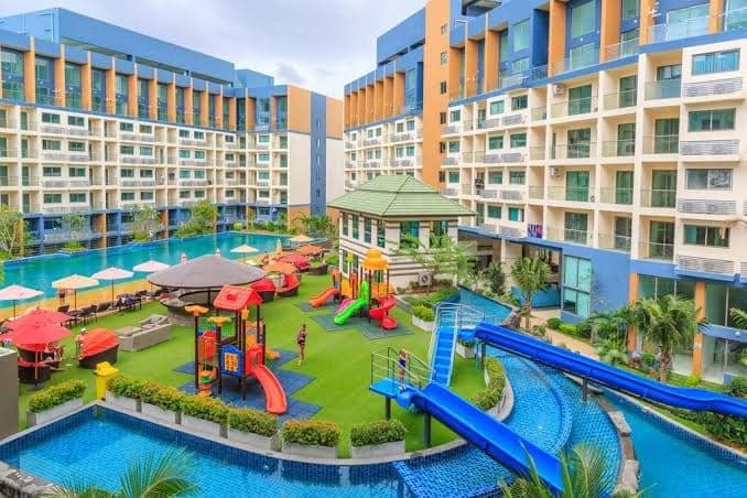 Family-friendly resort pool area with blue water slides, a colorful playground, loungers and umbrellas, surrounded by orange and blue buildings.