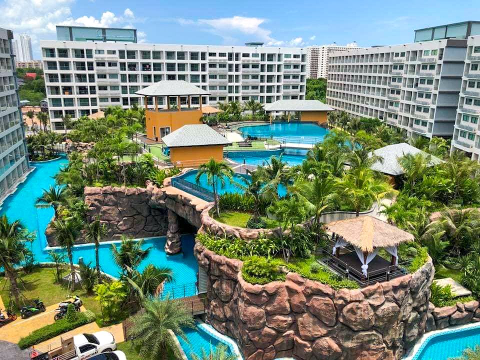 A tropical resort with winding blue pools rock arches palm trees and a central yellow pavilion surrounded by white apartment buildings