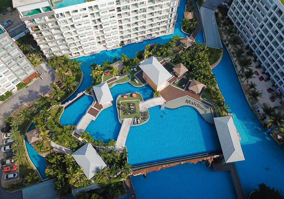 Aerial view of a resort pool complex with bright blue water weaving between palm trees and white cabanas, labeled Maldives on a curved walkway between high-rise buildings.