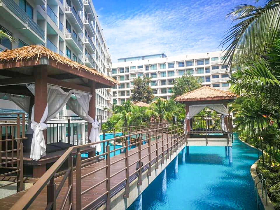 Resort-style pool area with thatched-roof cabanas along a winding blue pool, palm trees and white apartment buildings in the background.
