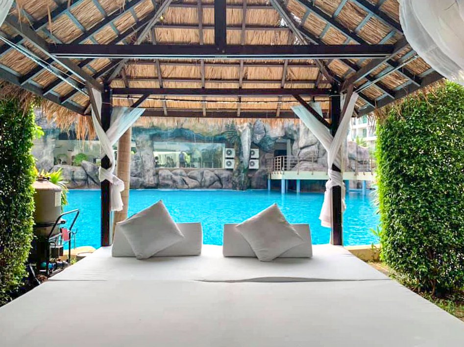 Poolside cabana with a white daybed and cushions facing a bright blue swimming pool at a tropical resort.