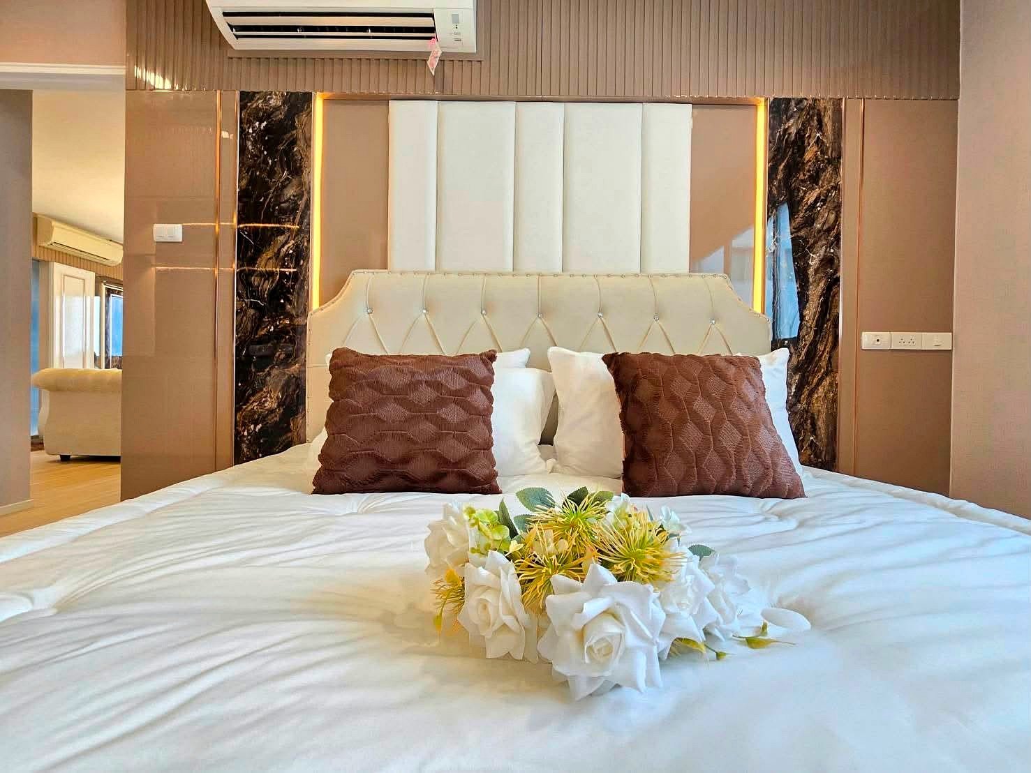 Lumpin_NorthPattaya_Sukhumvit_Dlc643_11 Luxurious bedroom scene with a tufted cream headboard, white sheets, and two brown textured pillows with a floral bouquet in the center of the bed.
