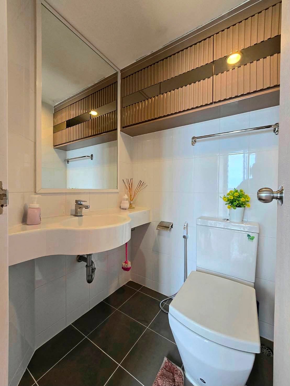 Lumpin_NorthPattaya_Sukhumvit_Dlc643_21 Bright modern bathroom with a white sink, large mirror, and wall cabinet above; toilet to the right and a small plant on the tank.