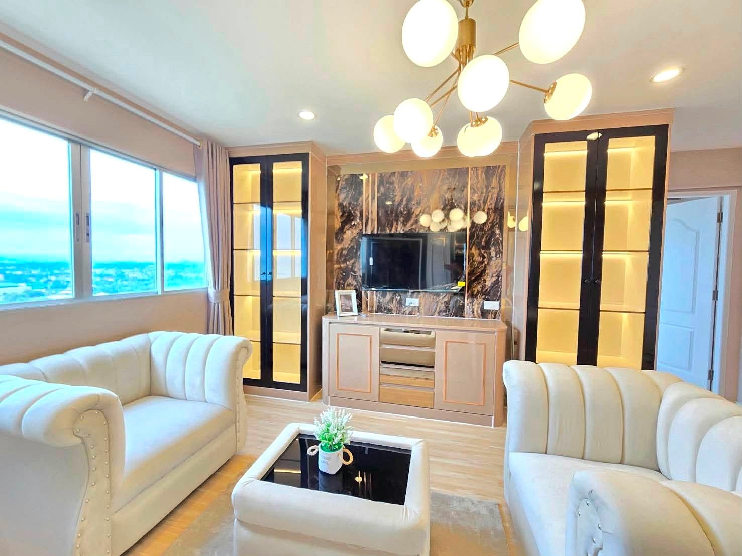 Lumpin_NorthPattaya_Sukhumvit_Dlc643_23 Bright living room with cream tufted sofas, a marble feature wall around the TV, and tall glass-front cabinets with warm backlighting beside a large window view of the city/landscape.
