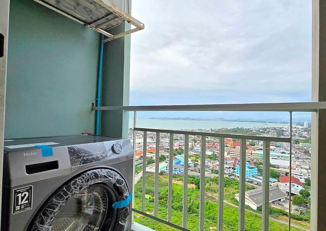 Balcony laundry area with a front-loading washing machine covered in plastic, beside an outdoor air conditioner and rusted bracket, overlooking a coastal city and sea.