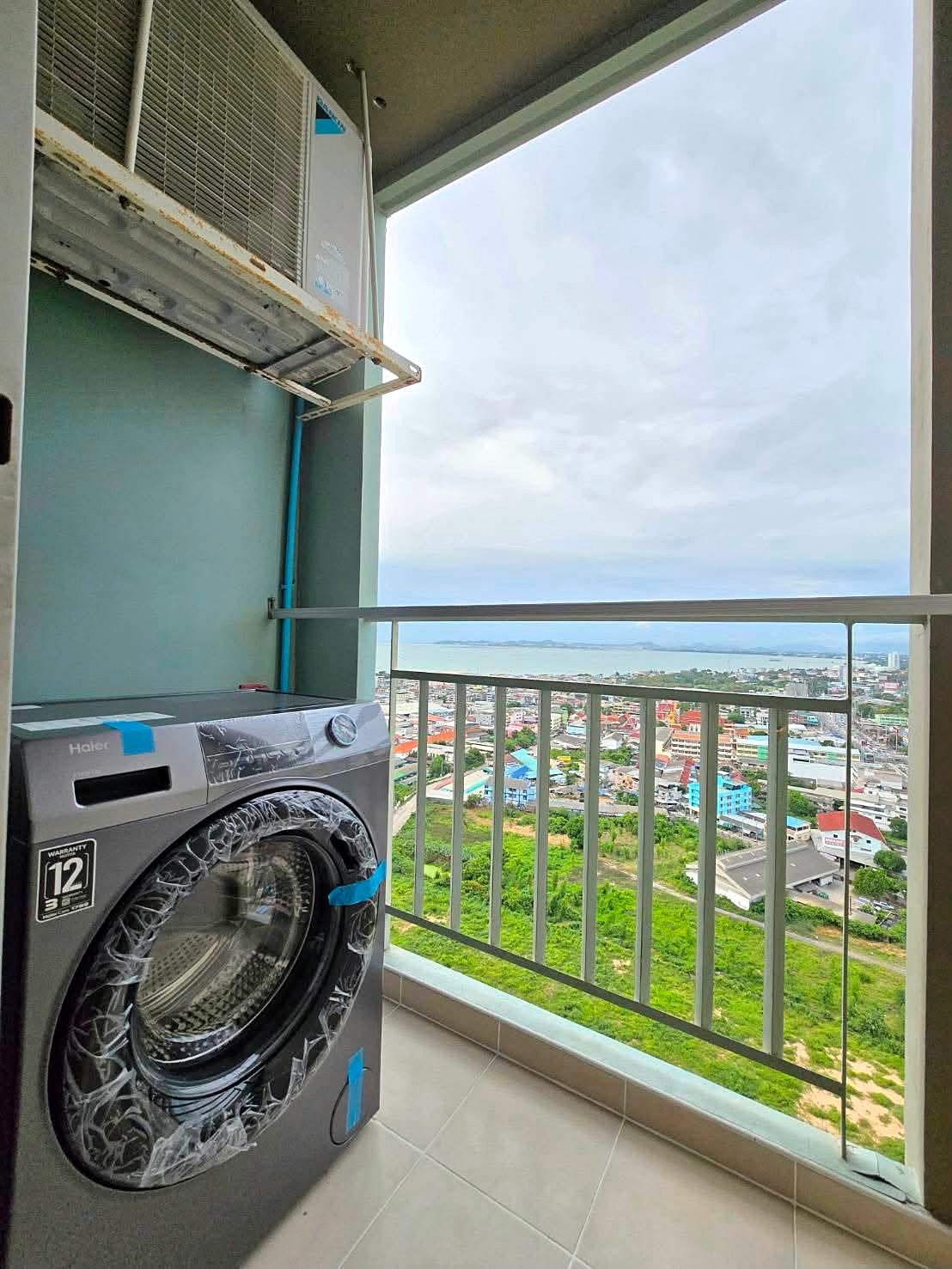 Lumpin_NorthPattaya_Sukhumvit_Dlc643_25 Balcony laundry area with a front-loading washing machine covered in plastic, beside an outdoor air conditioner and rusted bracket, overlooking a coastal city and sea.