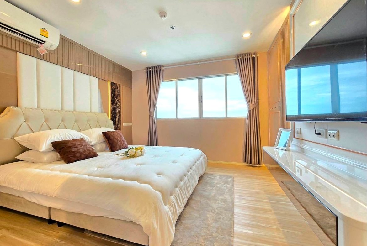 Luxurious hotel bedroom with a large tufted bed, white linens, and brown throw pillows, facing a window with drapes and a wall-mounted TV on the right.