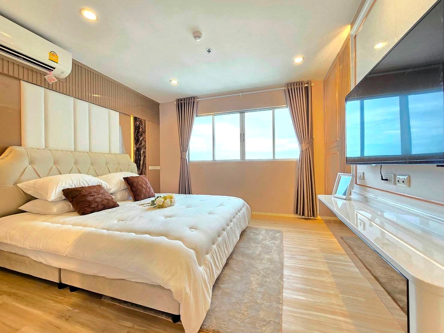 Lumpin_NorthPattaya_Sukhumvit_Dlc643_3 Luxurious hotel bedroom with a large tufted bed, white linens, and brown throw pillows, facing a window with drapes and a wall-mounted TV on the right.