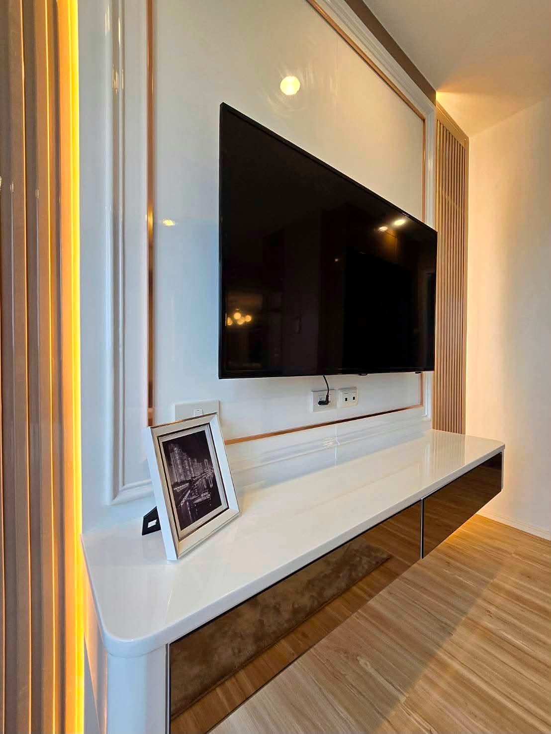 Lumpin_NorthPattaya_Sukhumvit_Dlc643_6 Modern living room entertainment wall with a mounted flat-screen TV above a glossy white console and a framed photo, accented by warm vertical LED lighting.