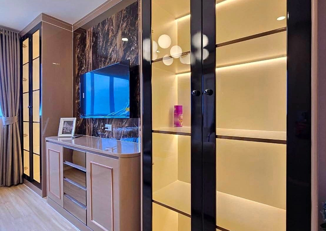 Modern living room corner with tall glass-front display cabinets lit from within, a wall-mounted flat-screen TV, and a light wood sideboard beneath on a marble-accent wall.
