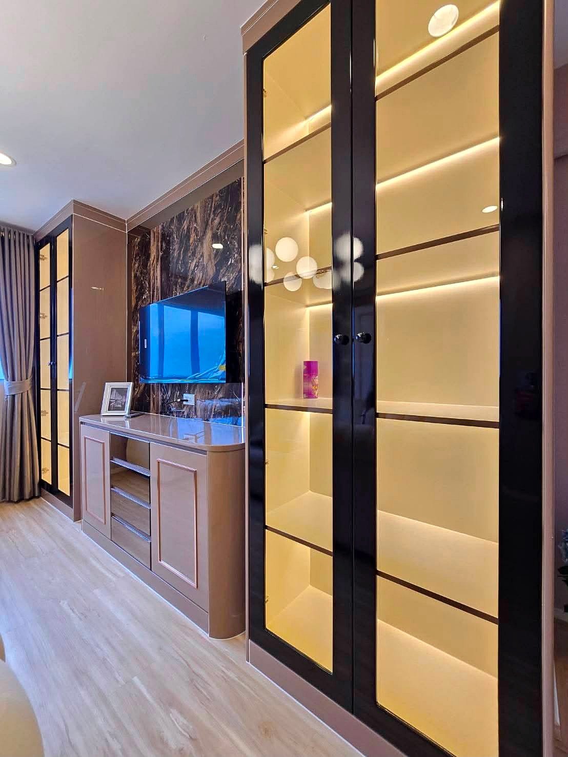 Lumpin_NorthPattaya_Sukhumvit_Dlc643_8 Modern living room corner with tall glass-front display cabinets lit from within, a wall-mounted flat-screen TV, and a light wood sideboard beneath on a marble-accent wall.