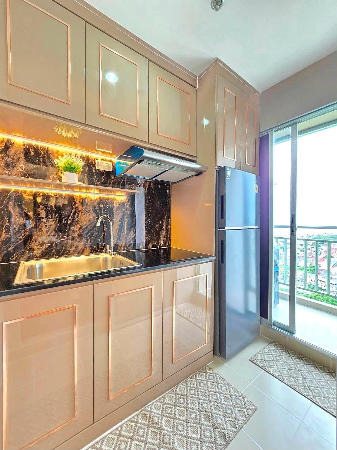 Lumpin_NorthPattaya_Sukhumvit_Dlc643_9 Modern kitchen with beige glossy cabinets, black marble backsplash, and stainless steel sink with a chrome faucet beside a sliding balcony door.